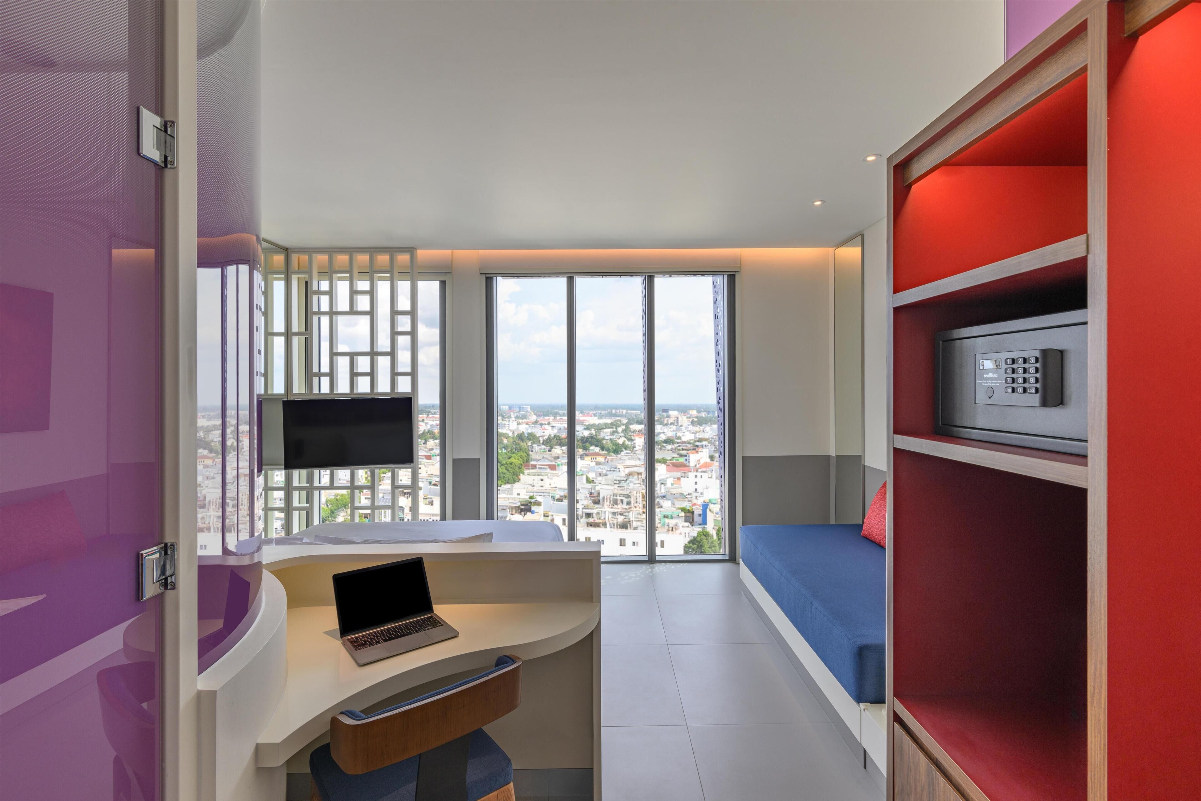 Una habitación de hotel moderna con un escritorio curvo, una laptop, un ventanal con vistas a la ciudad, un estante rojo y un sofá azul.