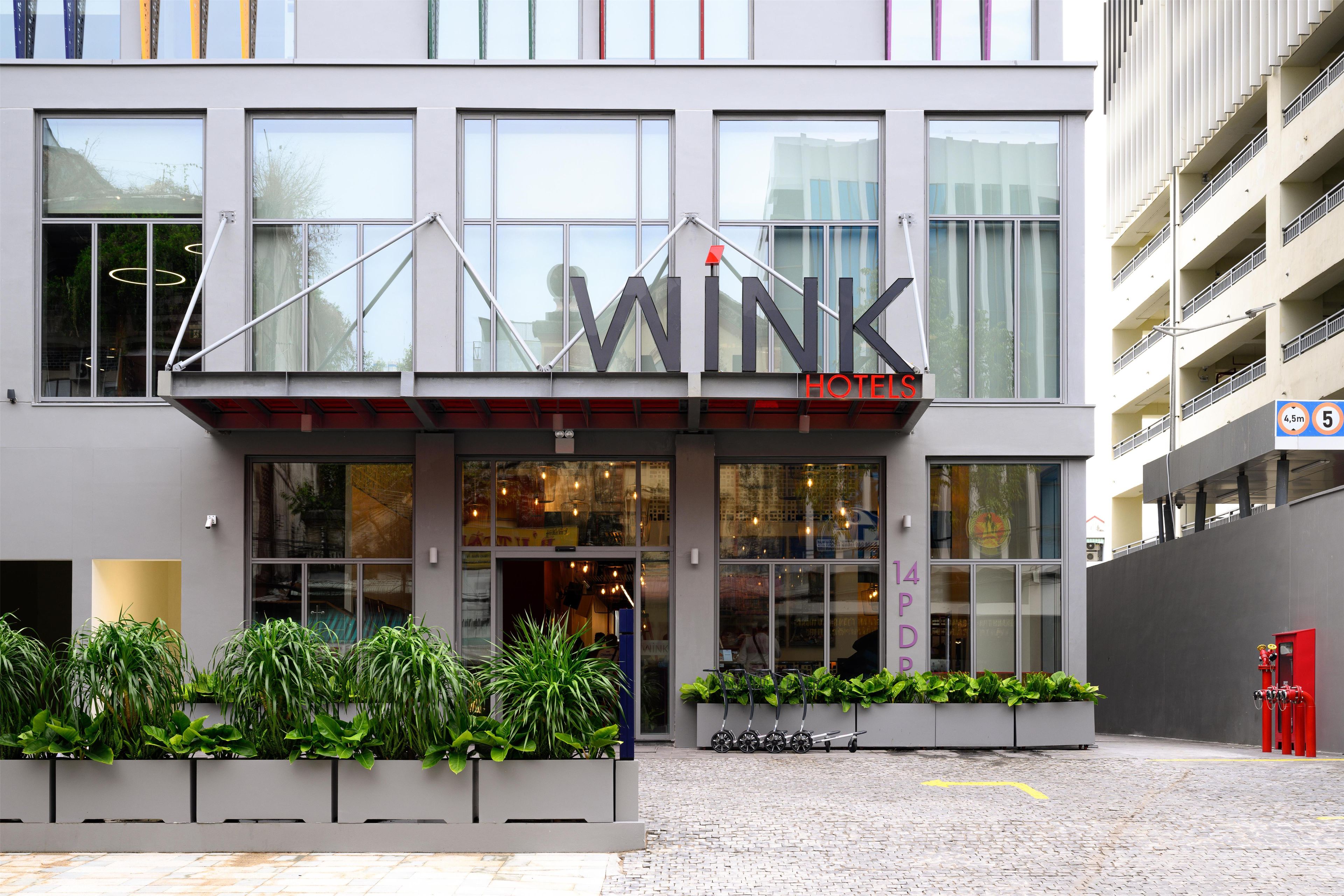 A modern hotel with a prominent 'WINK' sign, large windows, and a paved entrance area with planters.