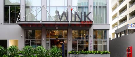 A modern hotel with a prominent 'WINK' sign, large windows, and a paved entrance area with planters.