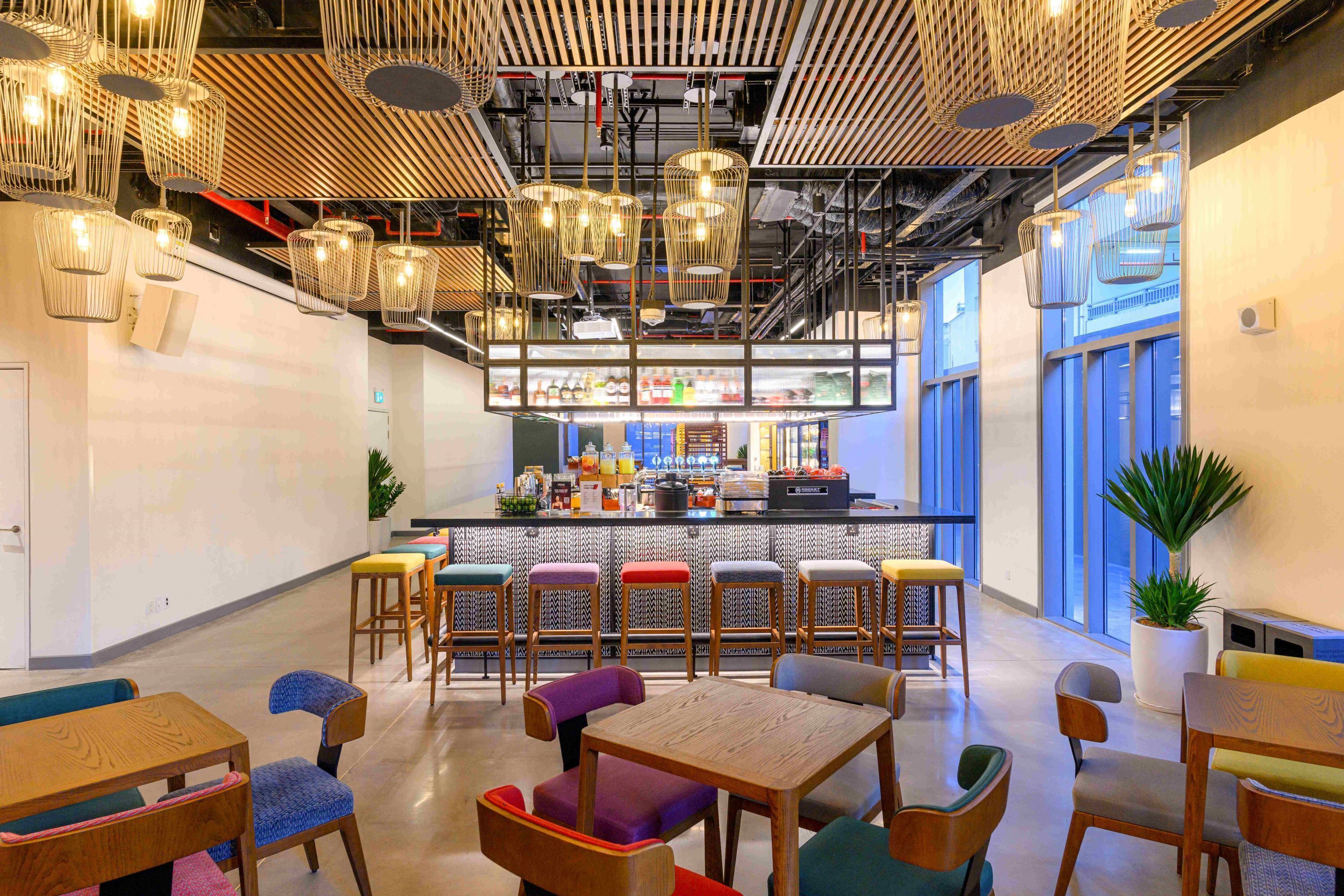 Un café moderno con mesas de madera y sillas coloridas, una zona de bar y grandes ventanales.