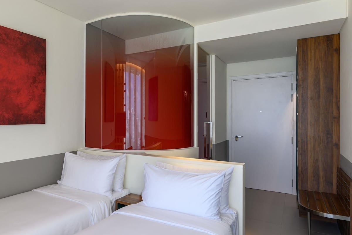 Una habitación de hotel con una cama, una mesita de noche, un espejo, un cuadro abstracto rojo y un armario de madera.