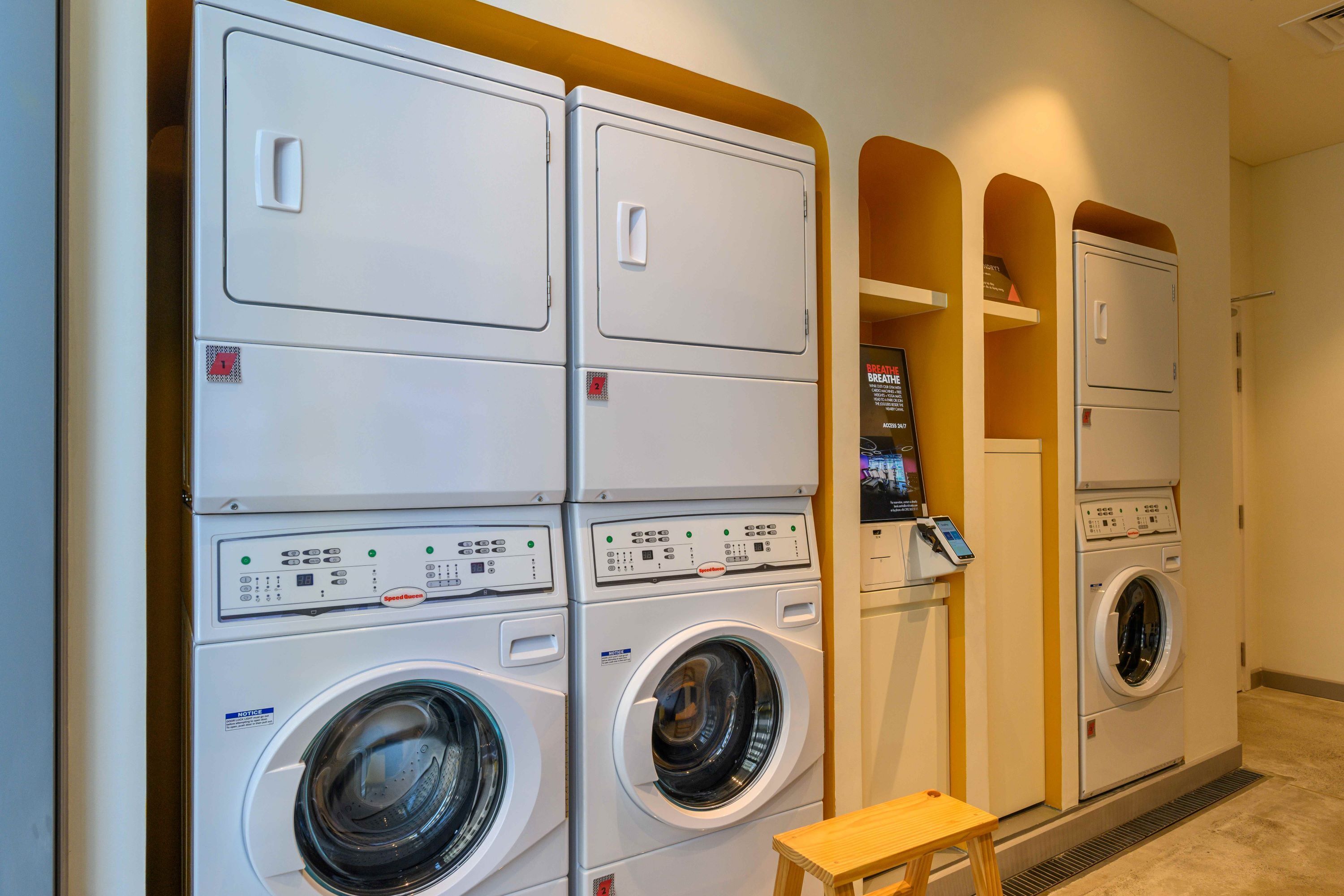 A laundry facility with multiple stacked washing machines and dryers, a control panel, and a small wooden stool.