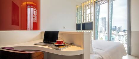 A modern hotel room with a desk, laptop, and a cup, a bed, and a large window with a city view.