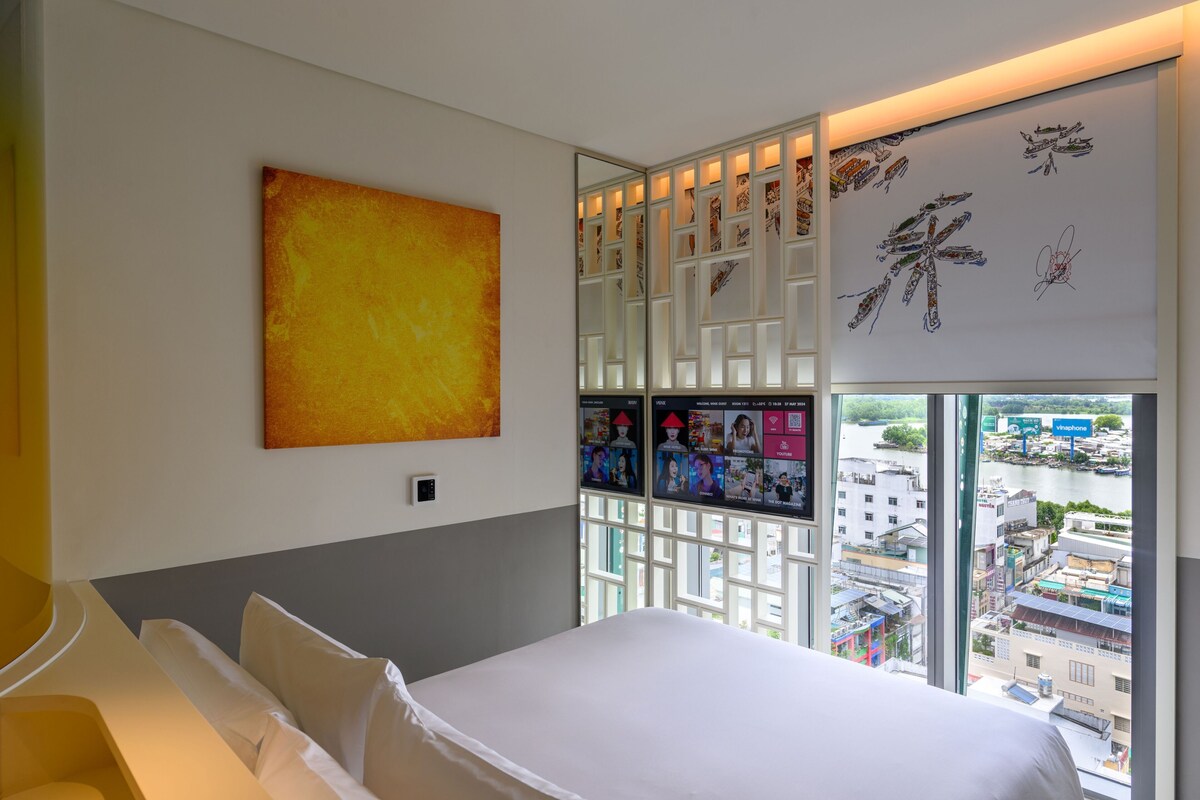 Habitación de hotel con cama, un ventanal grande, vista a la ciudad, una pared con obras de arte y una separación con fotografías enmarcadas.
