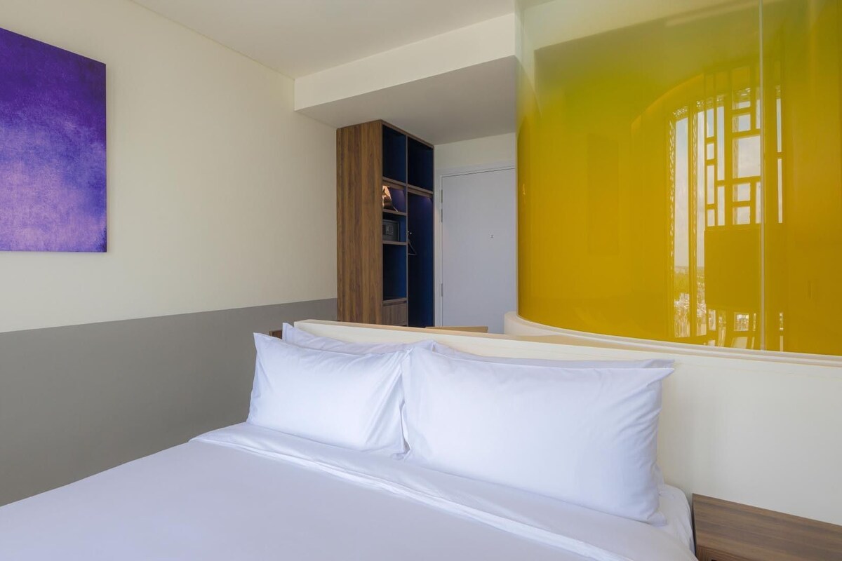 Una habitación de hotel con una cama, un cuadro abstracto púrpura y vistas a un edificio amarillo.