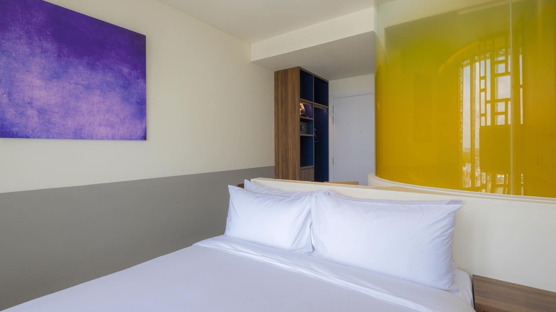 A hotel room with a bed, a purple abstract painting, and a view of a yellow building.