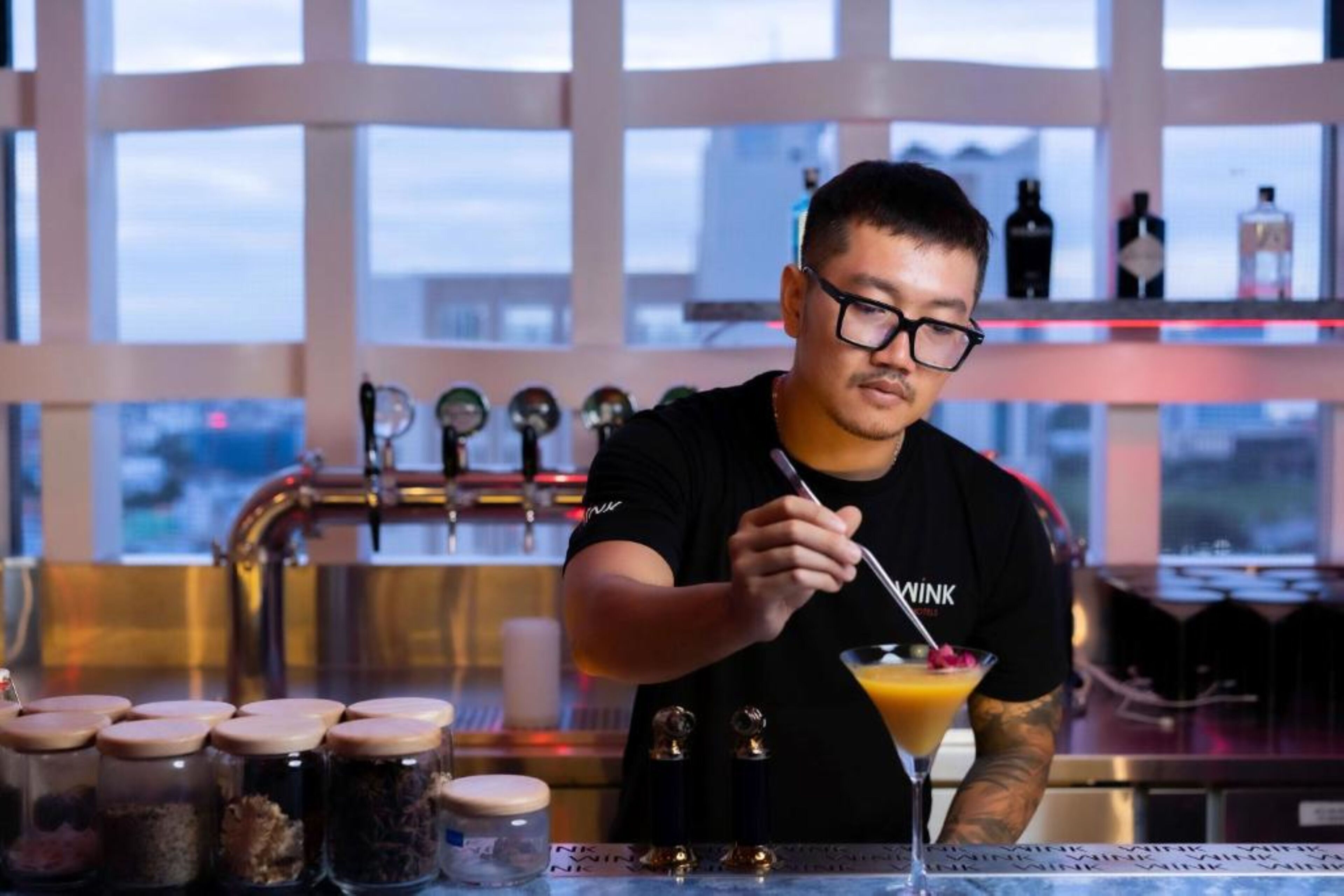 A bartender preparing a drink at a bar with a city view, shelves with bottles, and jars of ingredients.