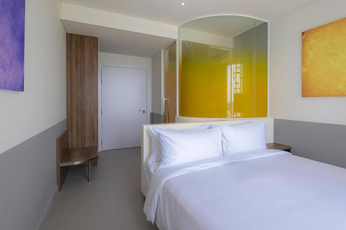 A hotel room with a bed, a wooden bench, a white door, and a large mirror.