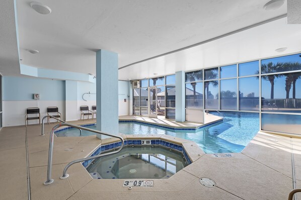 An indoor swimming pool with a hot tub, clear water, and a view of palm trees outside.