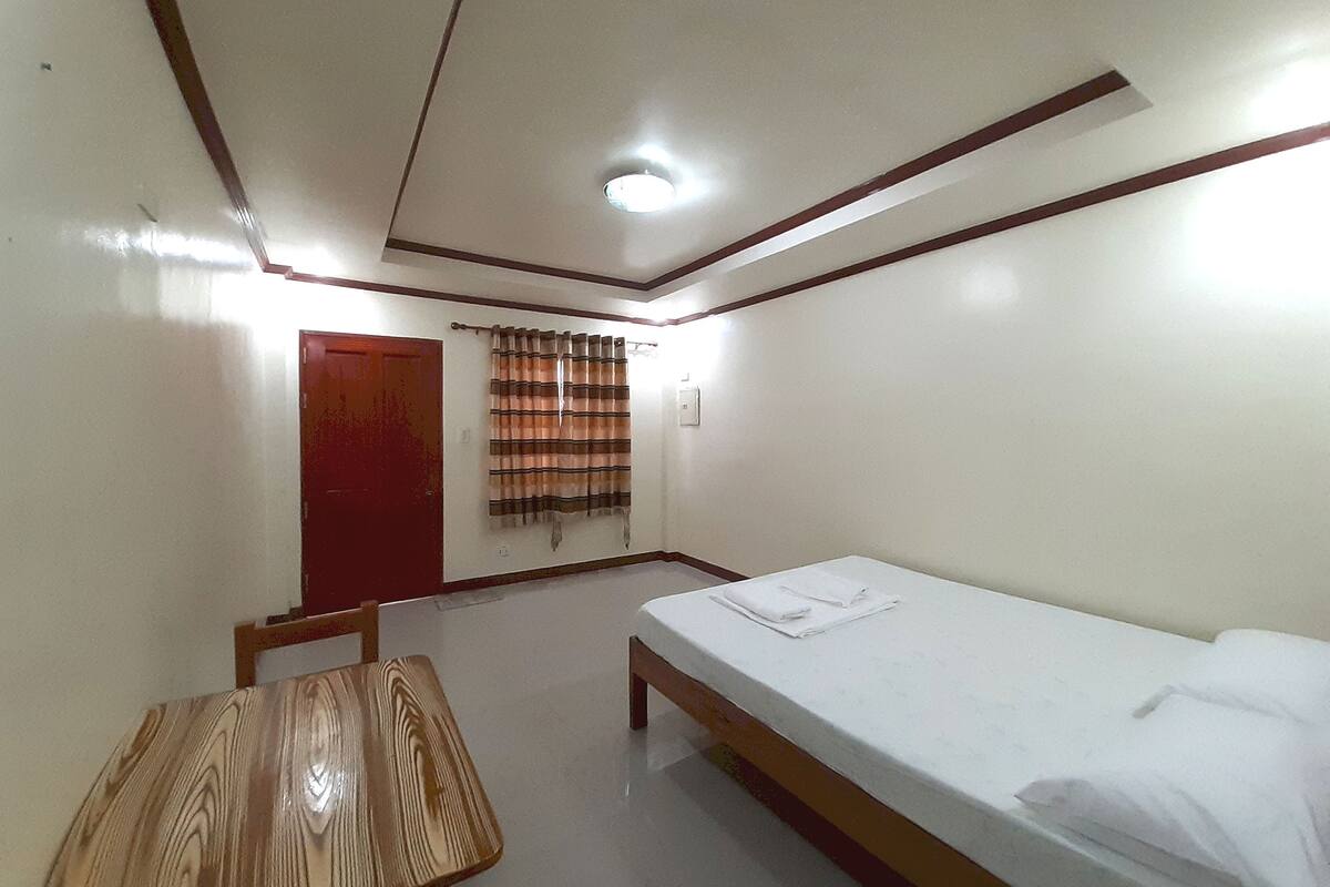 A room with a single bed, a wooden table, a red door, and a curtain.