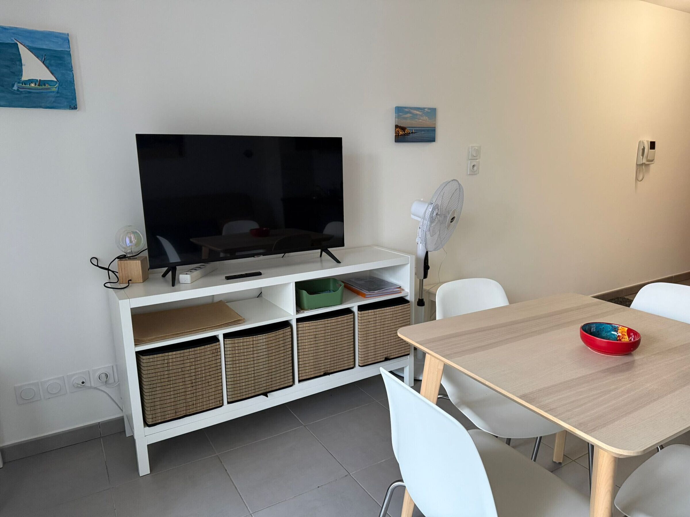 A modern living room with a flat-screen TV, white furniture, a dining table, and a small fan.