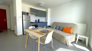 En kompakt opholdsstue med tekøkken, spiseplads med bord og stole, samt en sofa med puder.