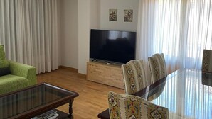 A living room with a flat-screen TV, a glass coffee table, a sofa, patterned chairs, and a wooden cabinet.
