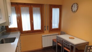 A compact kitchen with a dining table, chairs, a clock, and a window with curtains.