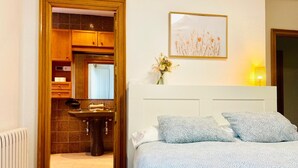 Un dormitorio con cama, almohadas, una mesita de noche, un cuadro en la pared y un baño visible a través de una puerta abierta.