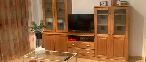 A living room with a TV, wooden cabinets, a glass coffee table, and a plant.
