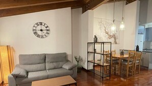 Un salon avec un canapé gris, une table basse en bois, une horloge noire, un coin repas avec une table et des chaises en bois, et une cuisine à l’arrière-plan.