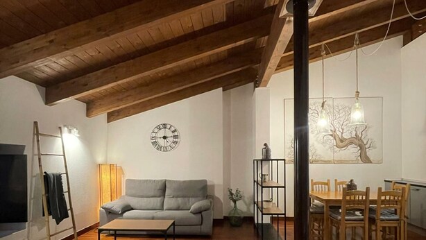 Un salon chaleureux avec un plafond en bois, un canapé gris, une table basse et un coin repas comprenant des chaises et une table en bois.