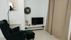 Una sala de estar con un sofá verde oscuro, un televisor sobre un mueble blanco, una lámpara de pie, un adorno circular decorativo y una cortina color crema.