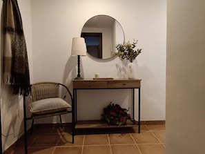 A room with a chair, a table with a mirror, a lamp, and a vase with flowers.
