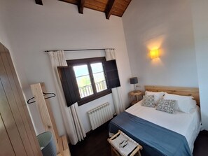 A bedroom with a bed, a window with curtains, a bedside lamp, a wooden headboard, and a small chair.