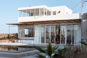 A modern two-story house with a pool, large glass windows, and a stone wall. - An architectural oasis with large swimming pool, along a breath-taking scenery. (Lajares)