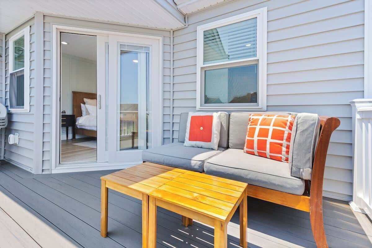 Eine überdachte Veranda mit einem Holztisch, einem grauen Sofa mit orangefarbenen und weißen Kissen und einer Schiebetür zu einem Schlafzimmer.