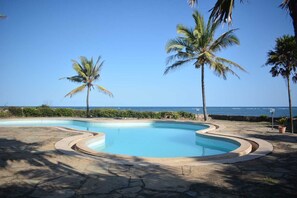 Ein Pool mit geschwungener Kante, umgeben von Steinfliesen, Palmen und Blick auf den Ozean.