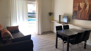 A modern living room with a sofa, a dining table with chairs, a TV on a stand, and a painting on the wall. - Denia: Appartement 1 Chambre avec Piscine, Clim et Parking à 500m de la Plage (Denia)