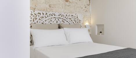 A bed with a white headboard and a grey blanket, set against a textured stone wall.