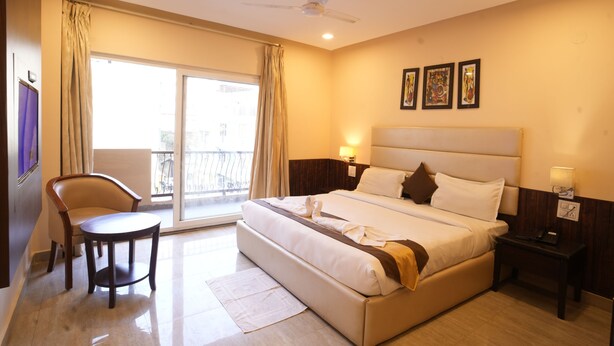 A hotel room with a bed, a chair, a desk, a ceiling fan, and a balcony with curtains. - Hotel Royale Hills (Narendranagar)