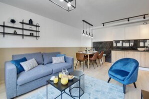 Un soggiorno moderno con divano, tavolino e poltrona blu. È presente una zona pranzo con tavolo e sedie. Si intravede la cucina, caratterizzata da un piano di lavoro e pensili neri.