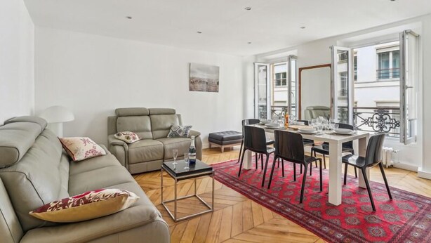 A modern living room with a sofa, a dining table set for four, and a red patterned rug. - 618 Suite Elisabeth - Stunning Apartment in Paris (Paris 10 Entrepôt)