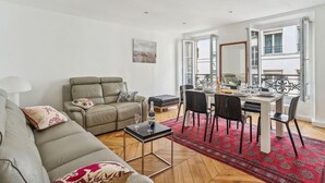 A modern living room with a sofa, a dining table set for four, and a red patterned rug. - 618 Suite Elisabeth - Stunning Apartment in Paris (Paris 10 Entrepôt)