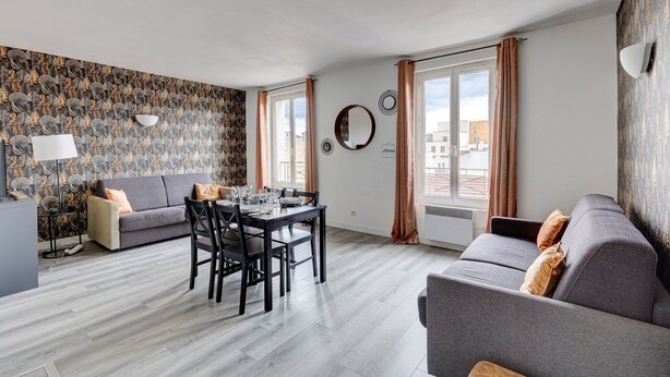 Een moderne woonkamer met een grijze bank, een eettafel met stoelen en behang met een patroon.