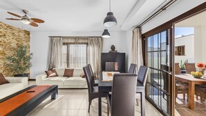 Un salon moderne avec un canapé, une table à manger et des chaises. On y trouve un ventilateur de plafond, une plante en pot et une fenêtre avec des rideaux.