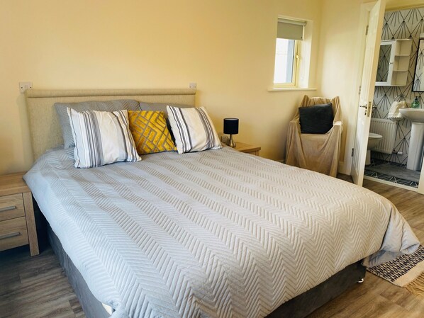 Una cama bien hecha con una colcha y almohadas estampadas, una mesita de noche con una lámpara, y vista a un baño con espejo y lavamanos.