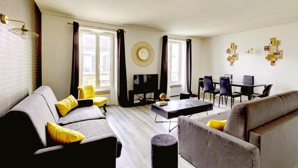 Un salon moderne avec un canapé, une table basse et un téléviseur. Les fenêtres sont dotées de rideaux, il y a un miroir rond et une œuvre d’art murale.