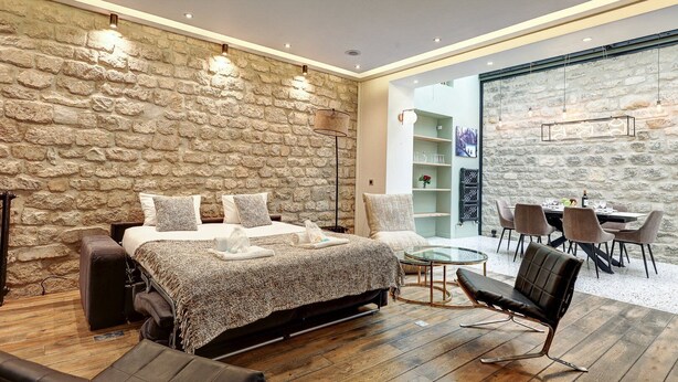 A bedroom with a stone wall, a bed with a patterned blanket, a glass table, and a seating area with chairs. - 334 Parisian Loft - Superb Apartment in Paris (Paris 10 Entrepôt)