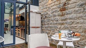 An outdoor seating area with wicker chairs and a table, a stone wall, and a glass door leading to a kitchen area. - 334 Parisian Loft - Superb Apartment in Paris (Paris 10 Entrepôt)