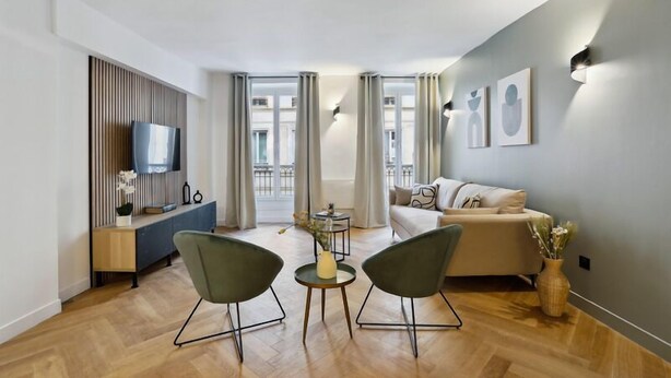 A modern living room with a sofa, two chairs, a TV, a small table with a vase, and a balcony with curtains. - 266 Suite Montefiore Superb apartment in Paris. (Paris 02 Bourse)
