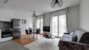 A modern kitchen with a built-in oven and refrigerator, a dining area with a table and chairs, a gray sofa, and a large window with a view of buildings. - 340 Suite Emile - Superb apartment (Clichy)