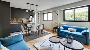 A modern living room with a blue sofa, a dining area with a table and chairs, and a kitchen with dark cabinets and a marble backsplash. - 217 Suite Trocadero - Superb apartment in Paris (Paris 16 Passy)