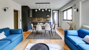 A modern living room with a blue sofa, a dining table with chairs, and a kitchen area with a dark cabinet and hanging lights. - 217 Suite Trocadero - Superb apartment in Paris (Paris 16 Passy)
