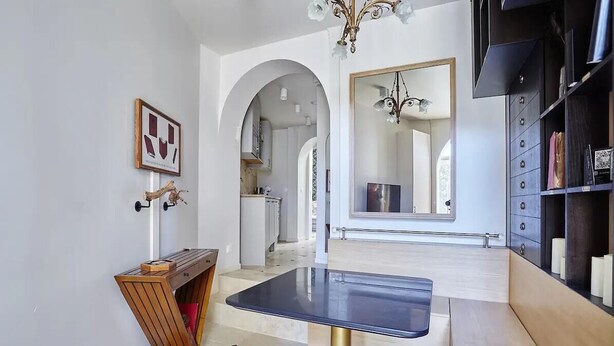 Un intérieur moderne avec une table centrale, une bibliothèque et une porte en arc donnant sur une cuisine.
