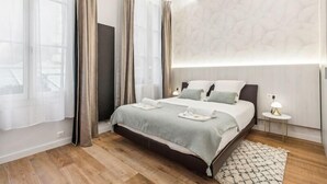 Une chambre à coucher moderne avec un grand lit, une table de chevet avec une lampe et une vue sur l’extérieur à travers des rideaux légers.