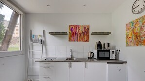 A modern kitchen with white cabinets, a stainless steel sink, a microwave, and a kettle. There are two paintings on the wall, a window with a view of buildings, and a clock. - 235 Home Madrid Concept (Paris 15 Vaugirard)