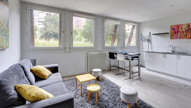 A modern living room with a grey sofa, a small coffee table, and a dining area with a table and chairs. Large windows provide natural light and views of the outside. - 235 Home Madrid Concept (Paris 15 Vaugirard)