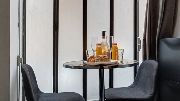 Una pequeña mesa redonda con un croissant, dos vasos y dos botellas de zumo de naranja y vino, frente a una pared blanca con rayas verticales negras.