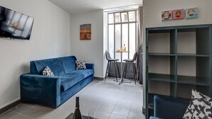 Una sala de estar con un sofá azul, un televisor montado en la pared, un cuadro y una ventana con cortinas.